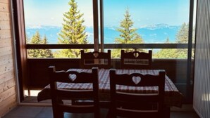 Un interno in legno con tavolo e sedie, ampie finestre che offrono vista su alberi e montagne.