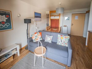 En hyggelig stue med en grå sofa, en lille træstol, et hvidt sengebord og et billede i ramme på væggen.