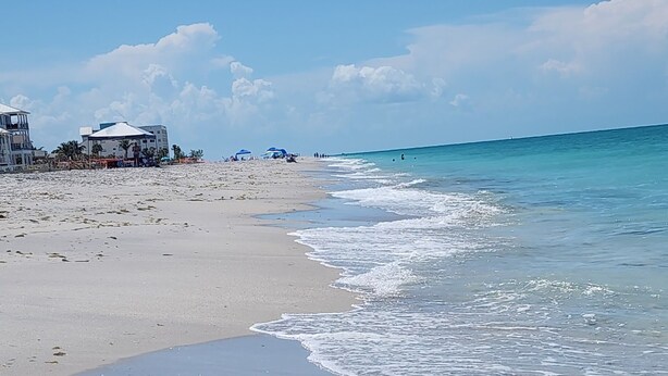 Una playa con agua cristalina, una orilla arenosa y edificios al fondo.