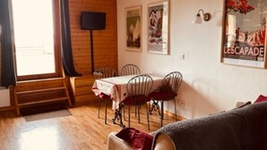 A room with a wooden floor, a sofa, a dining table with chairs, a TV mounted on the wall, and a poster on the wall. - Apartment 'Les Edelweiss 201' Large Terrace with Mountain and Valley Views (Chamrousse)