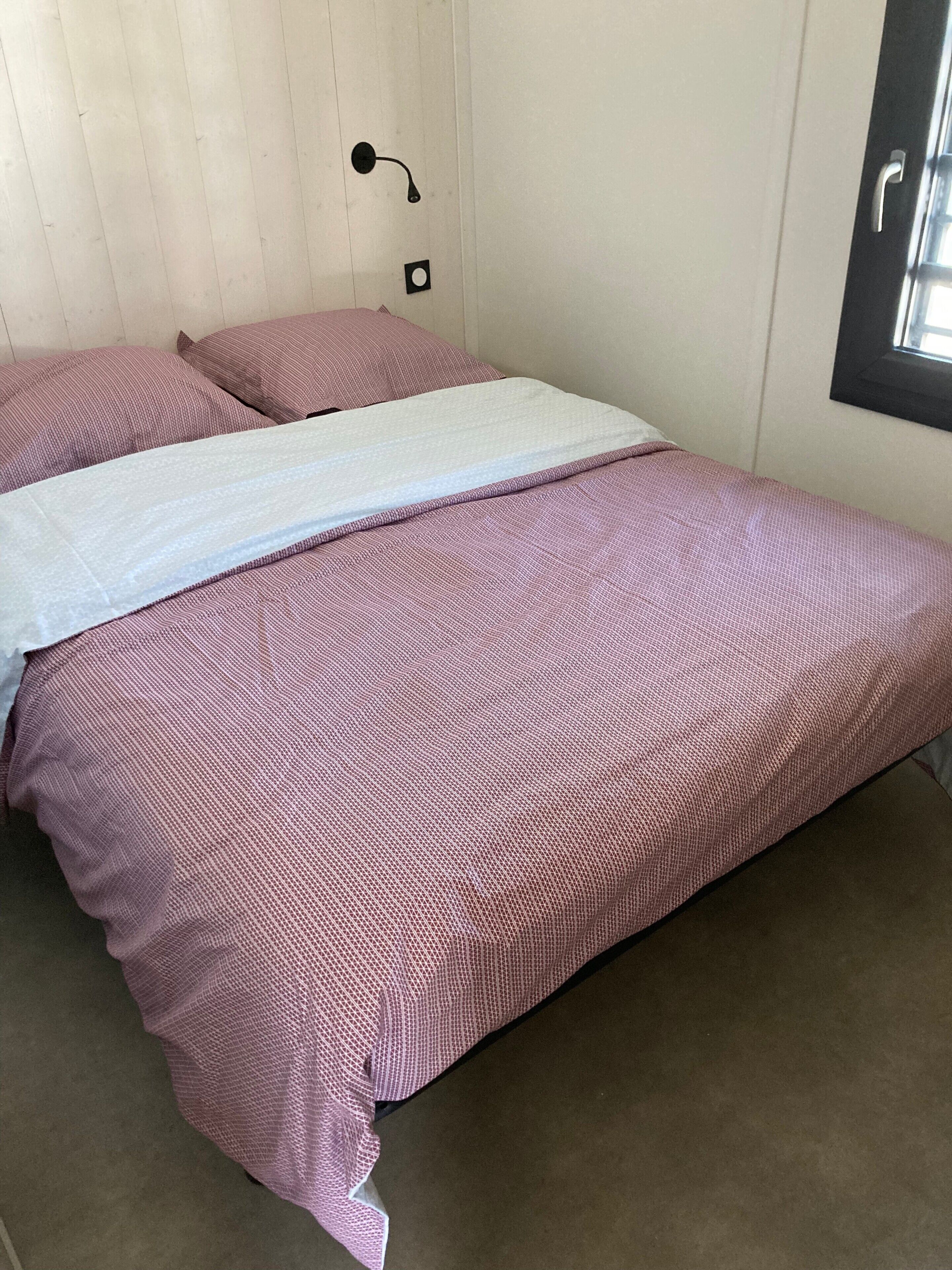 A bedroom with a bed, pillows, a wall-mounted light, and a window.