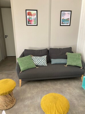 Ein graues Sofa mit gemusterten Kissen, zwei gerahmte Bilder an der Wand und zwei runde, gelbe Hocker auf dem Boden.