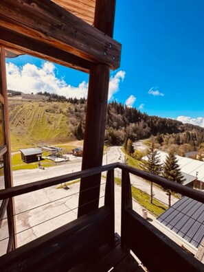 A wooden balcony with a view of a hilly landscape, a road, and a building with a solar panel roof. - Studio 'Le Bouquetin 304' for 2 – Les Balcons du Recoin, Chamrousse 1650 (Chamrousse)
