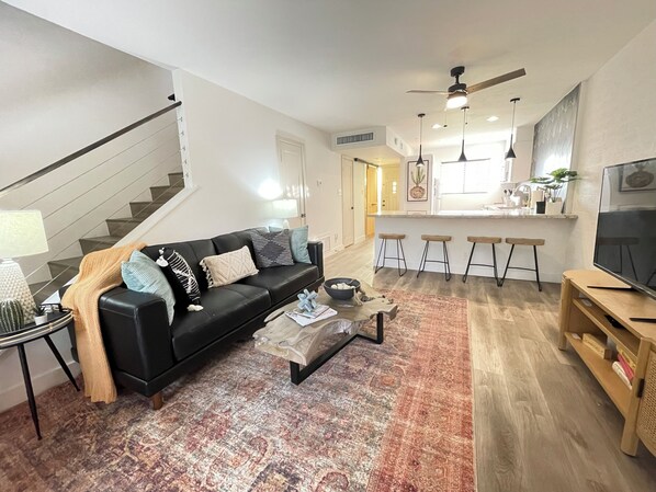 A modern living room with a black leather sofa, a coffee table, and a patterned rug. There is a kitchen area with bar stools and a large TV. - Southwest Meets Boho Old Town Scottsdale Townhome (Scottsdale)