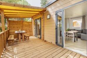 Eine Holzdielen-Terrasse mit Tisch und Bänken, einem überdachten Bereich und einer Glastür, die zu einem Innenraum mit einer Couch und einem Tisch führt.