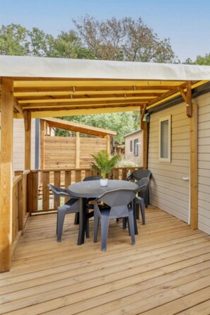 Eine Holzterrasse mit überdachter Veranda, einem Tisch mit Stühlen und einem kleinen Gebäude im Hintergrund.