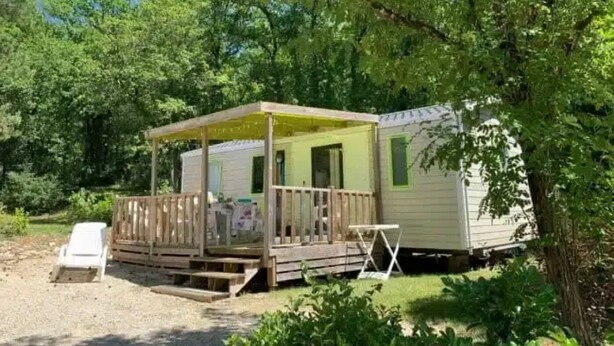 Una casa móvil con porche y una pequeña terraza, rodeada de árboles.