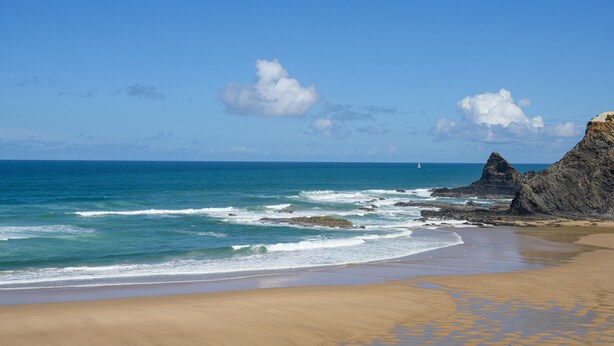 A beach with clear skies, waves, and a rocky outcrop.