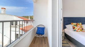 A balcony with a chair and a bed, a clear sky, and buildings in the background.