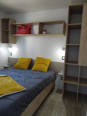 Ein Schlafzimmer mit einem Bett, zwei gelben Kissen, einer blauen Tagesdecke, einer roten Tasche auf einem Regal und einer Holzregaleinheit.