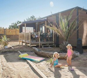 Ein Holzhaus mit einer Terrasse, einem Sandbereich mit Poolspielzeug und einer Palme.