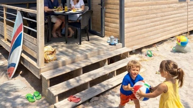 Een houten stacaravan met een overdekt terras, een surfplank en spelende kinderen.