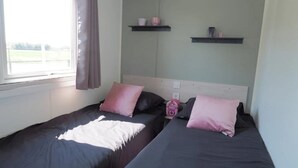Une chambre avec deux lits, une fenêtre avec des rideaux et deux étagères fixées au mur, sur lesquelles sont posés divers objets.