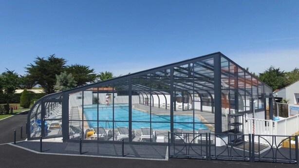Une piscine couverte, protégée par une structure en verre, entourée de chaises longues et d’une clôture blanche.