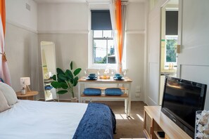 A bedroom with a bed, a bedside table with a lamp, a dresser, a mirror, a window with curtains, a TV, and a plant. - Boho Chic Studio Apartment, Next to CBD (Darlinghurst)