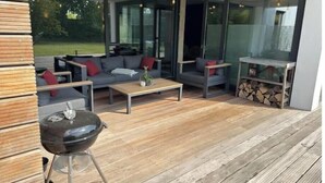Uma área externa moderna para relaxamento, com sofá, mesa de centro e churrasqueira, com portas de vidro e um deck de madeira.
