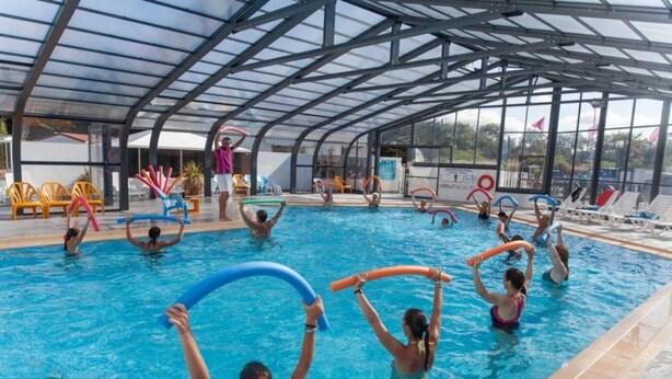 Ein Hallenbad mit Glasdecke, mehreren Bahnen und Liegestühlen am Beckenrand.