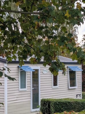 A single-story house with a beige exterior, a blue awning over the windows, and a small hedge in front. - Camping 2 étoiles - ccbg00a (Sémeries)