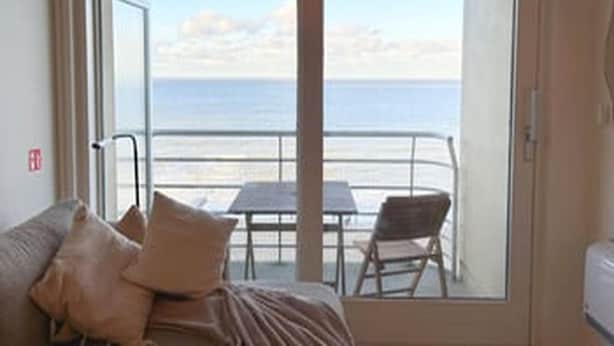 A room with a bed, a table, and a chair, with a view of the sea through large windows. - Stunning Seaview Apartment With Balcony (Blankenberge)