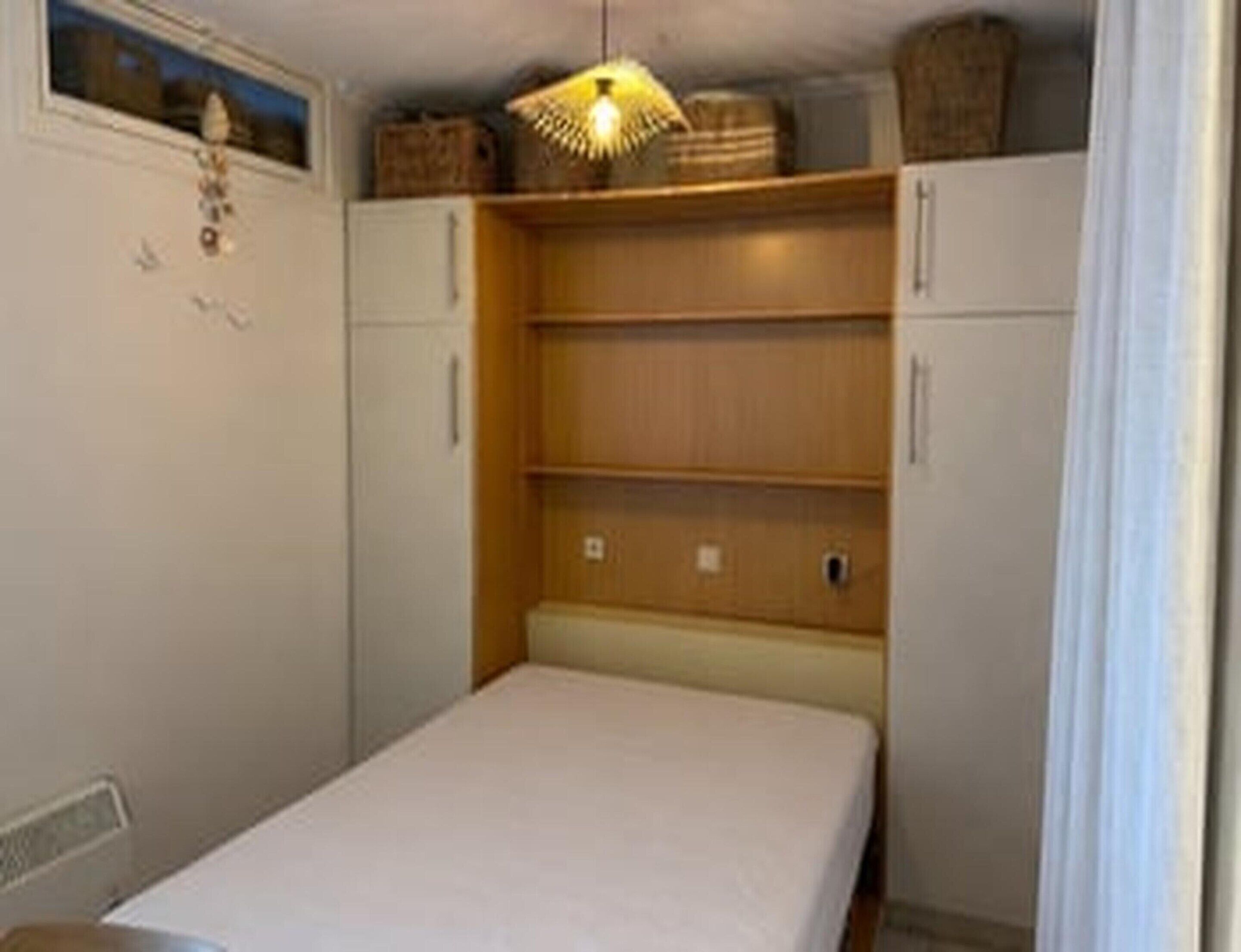 Una habitación pequeña y funcional con una cama individual, un estante de madera y una lámpara en el techo.