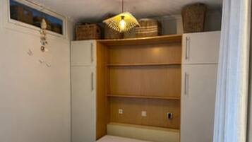 Una habitación pequeña y funcional con una cama individual, un estante de madera y una lámpara en el techo.