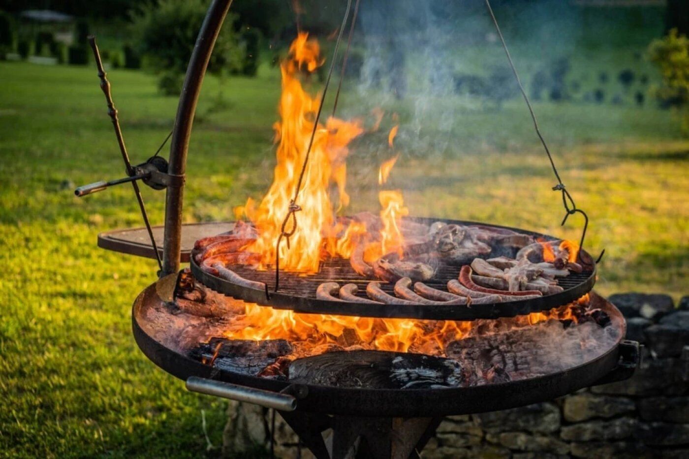 Un barbecue circulaire installé à l’extérieur, avec des saucisses en train de cuire sur une flamme nue, le tout entouré d’une zone herbeuse.