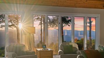 Una habitación con escritorio, dos sillas, lámpara y ventanas amplias con vista a árboles y montañas.