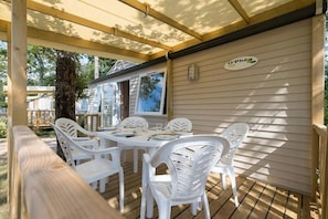 A covered outdoor area with a wooden deck, a round table with white plastic chairs, and a sign on the wall. - Camping 4 étoiles - Piscine  - ccbffd0 (Saint-Sulpice-de-Royan)
