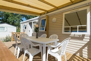 A covered outdoor patio with a table and chairs, a wooden deck, and a view of a neighboring house. - Camping 4 étoiles - Piscine  - ccbffda (Saint-Sulpice-de-Royan)