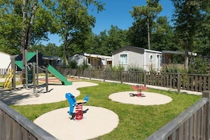 A playground with swings and a slide, surrounded by a wooden fence and houses in the background. - Camping 4 étoiles - Piscine  - ccbffda (Saint-Sulpice-de-Royan)