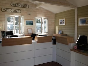 A reception desk with 'BIENVENUE' and 'WELCOME' signs, a window with a view of a house, and framed pictures on the wall. - Camping 4 étoiles - Piscine  - ccbffdb (Saint-Sulpice-de-Royan)