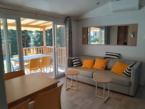 Ein modernes Wohnzimmer mit einer Couch, zwei kleinen Tischen, einem Esstisch und Stühlen. Es gibt einen Balkon mit Holzgeländer und Blick auf Bäume.