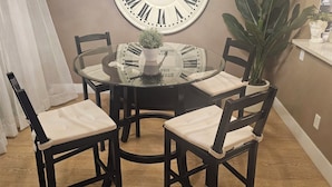 Een eetruimte met een ronde glazen tafel, vier stoelen, een potplant, een decoratieve klok en een vaas met plant.