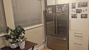 Een keuken met een roestvrijstalen koelkast, een raam met zonwering, een houten bankje met planten en een witte kast met lades.