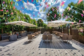 Eine Terrasse im Freien mit Holzboden, mehreren Tischen und Stühlen sowie bunten, dreieckigen Wimpeln.