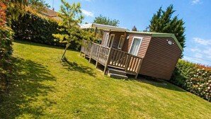 Eine kleine, eingeschossige Holzhütte mit einer umzäunten Terrasse, umgeben von einem gepflegten Rasen und Bäumen.