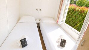 Ein kleines Zimmer mit zwei Betten, einem Fenster mit Blick auf Grünflächen und einer weißen Wand mit einem Regal.