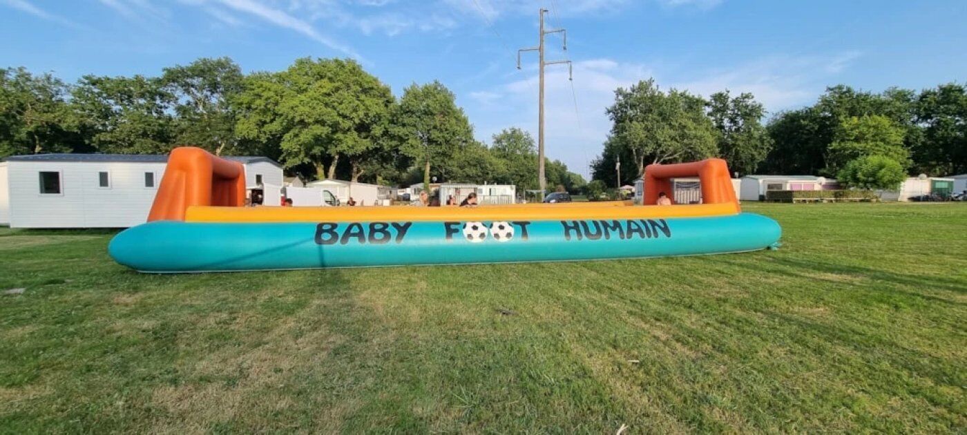 Una gran estructura inflable con las palabras "BABY FOOT HUMAN" sobre ella, ubicada en un campo cubierto de hierba con árboles y casas móviles al fondo.