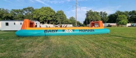 Una gran estructura inflable con las palabras "BABY FOOT HUMAN" sobre ella, ubicada en un campo cubierto de hierba con árboles y casas móviles al fondo.