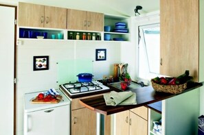 Eine kleine Küche mit Gasherd, Spüle und kleinem Kühlschrank. Über der Arbeitsfläche befinden sich Schränke und ein Fenster über der Spüle.