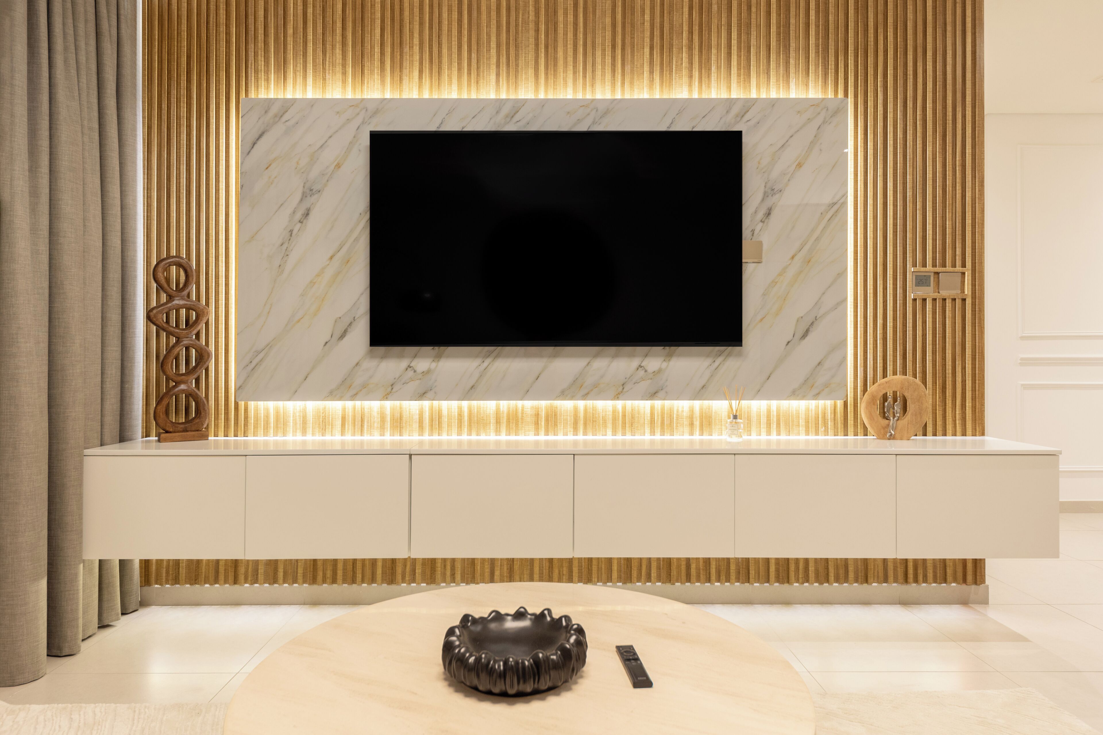 Una sala de estar moderna con un televisor de pantalla plana grande montado en una pared de mármol, una consola blanca elegante debajo y un cuenco decorativo sobre una mesa de centro redonda.