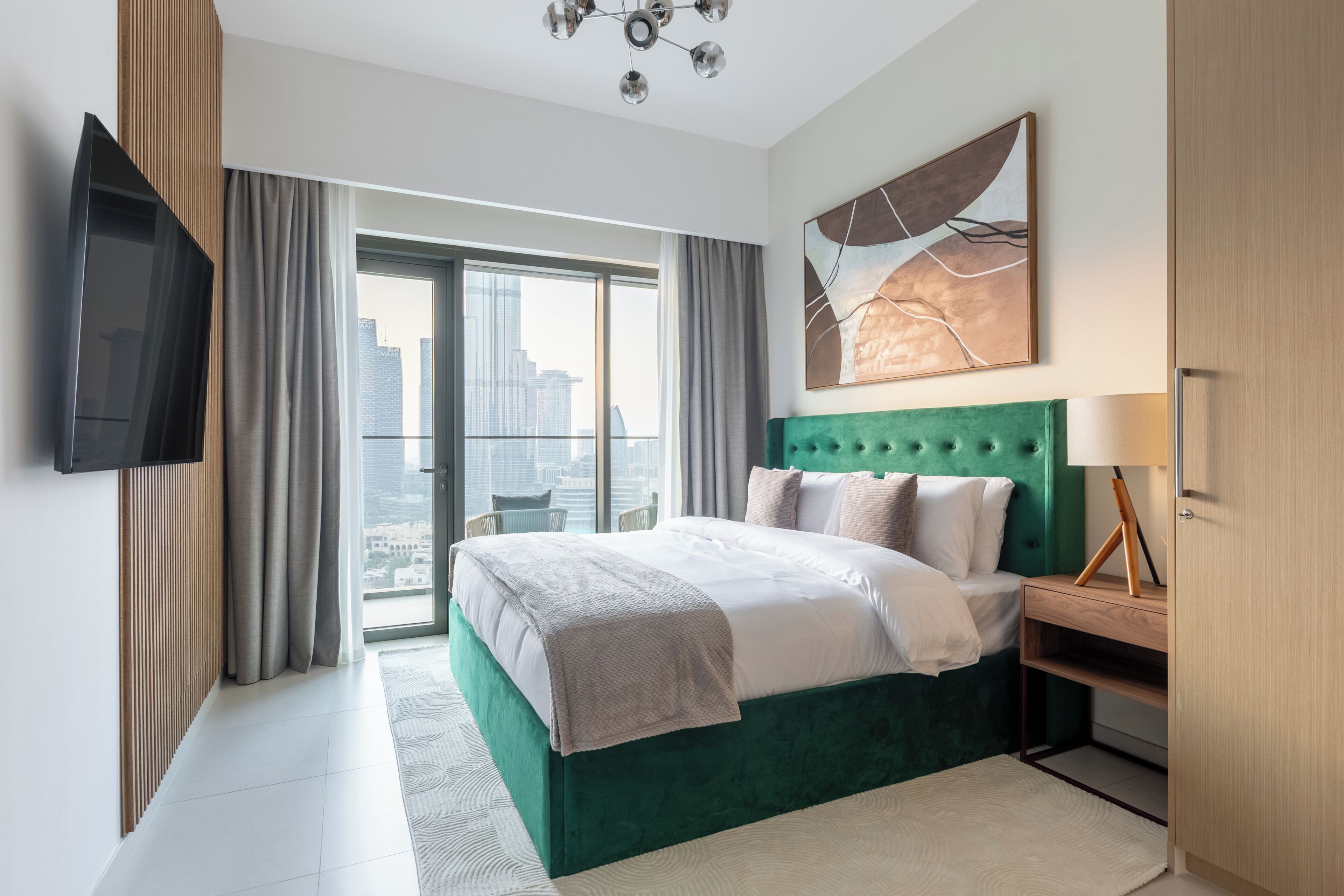 A modern hotel room with a large bed, a flat-screen TV, a city view, and a green upholstered headboard.