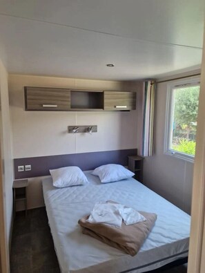 A hotel room with a bed, a bedside table, a wall-mounted shelf, and a window with curtains. - Camping 4 étoiles - Piscine  - ccbgaab (Mandelieu-la-Napoule)