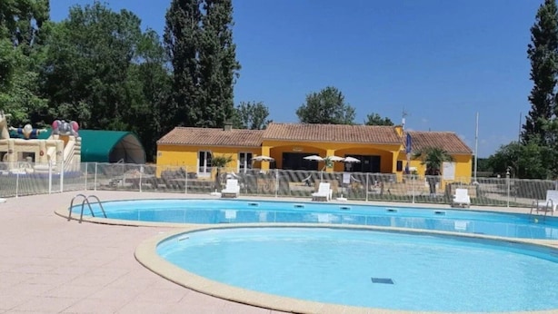 Ein Schwimmbad mit umzäuntem Bereich, ein gelbes Gebäude mit einem Ziegeldach und Bäume im Hintergrund.