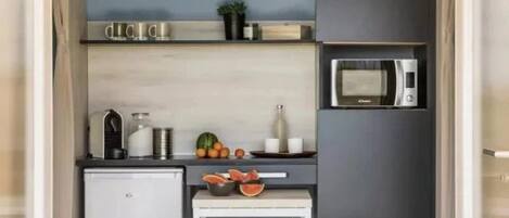 Una kitchenette compacta con microondas, un refrigerador pequeño, un lavamanos y una encimera con frutas y un tazón. Hay dos taburetes y una mesita.