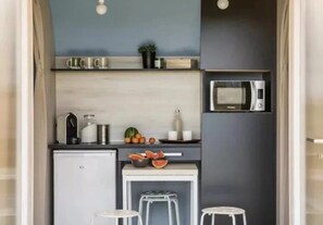 A compact kitchenette with a microwave, a small refrigerator, a sink, and a countertop with fruits and a bowl. There are two stools and a small table.