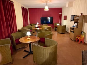 A room with wicker chairs, small round tables, a television, and a red curtain. - Camping 3 étoiles - Piscine  - ccbfig0 (Saint-Fort-sur-Gironde)