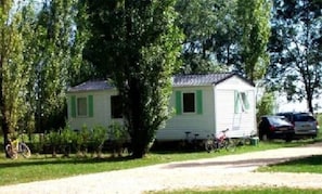 Una casa móvil de una sola planta, pequeña, con techo inclinado, contraventanas verdes y un porche delantero. Hay una bicicleta estacionada afuera y un coche en la entrada.