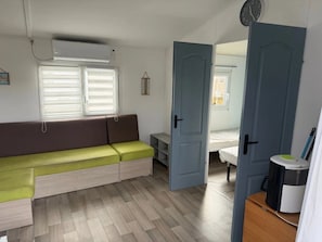 A compact room with a sofa, a bed, a door, and a small table with a trash bin. - Camping 3 étoiles - Piscine  - ccbfigc (Saint-Fort-sur-Gironde)