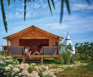 Eine Holzhütte mit einer großen, offenen Veranda, ein Tipi im Hintergrund und ein Blumengarten.